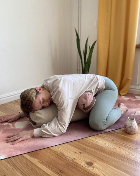 woman practising yoga on a mat lying on a bolster and a small buckwheat roller under her belly