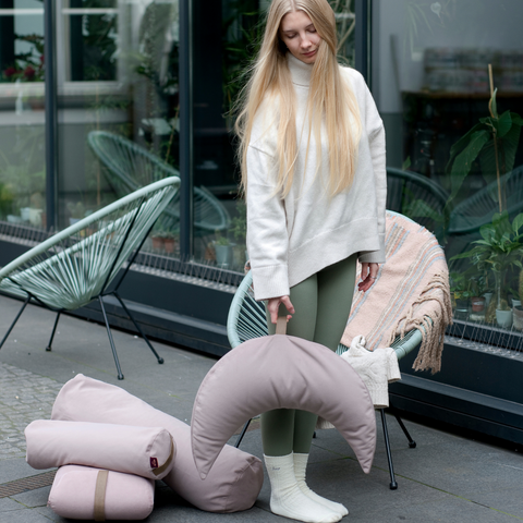 a woman on the terrace holds a pink moon pillow in her hand and other relaxation pillows from the same collection lie on the ground