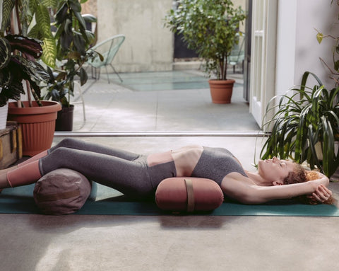 a woman practising yoga using a dark pink envelope pillow placed under her loins with her legs held on a bolster