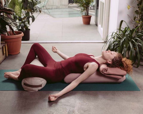 woman practising yoga on a mat with an enveloping pillow along her back and a bolster placed under her knees