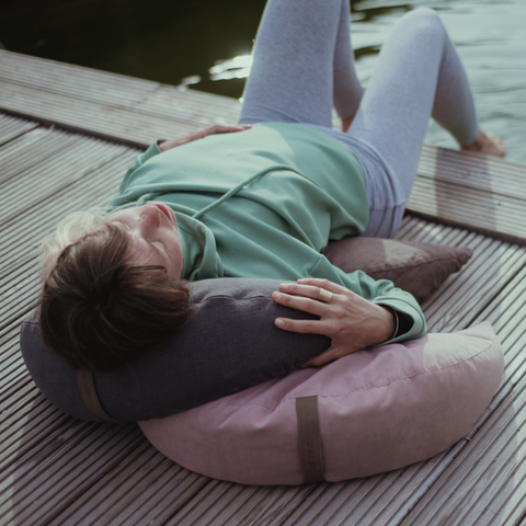 a woman lies on a pier by the lake resting her head and body on moon pillows from the be natural collection in linen and cotton
