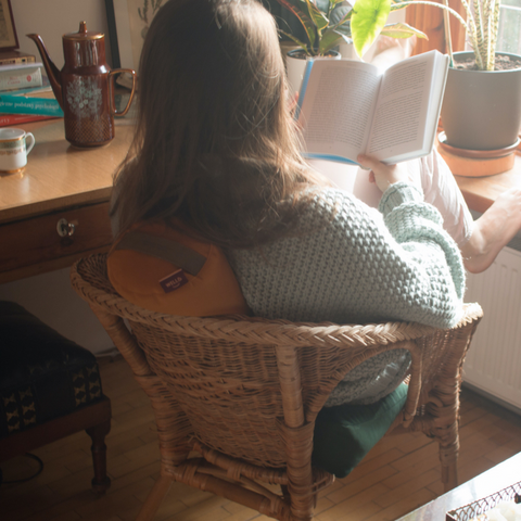 woman reading a book on a wicker armchair leaning her back against a yellow small buckwheat roller