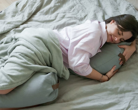 a woman who sleeps with a smile on her face cuddling up to a grey moon pillow with a bolster between her legs