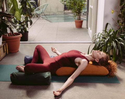 a woman practising yoga lying on a mat, supporting her back against a yellow envelope pillow and her knees against small buckwheat rollers
