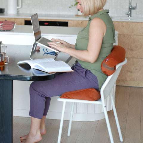 a woman working on a laptop using an orange wedge cushion to sit on a chair and resting her back on a small roller 
