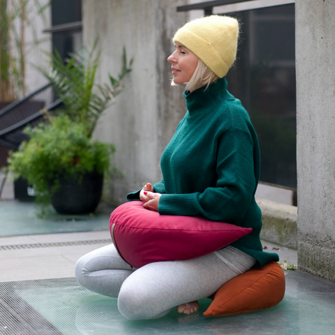 woman meditates sitting on orange moon cushion holding hands on pink moon cushion